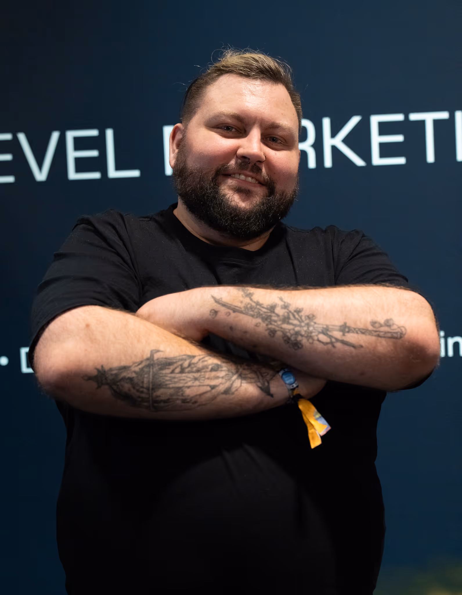 Portraitfoto von Patrik Garten, mit einem Lächelnden Gesicht, verschränkten Armen und sichtbaren Tattoos vor dunklem Messe-Hintergrund.